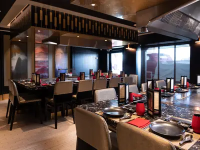 Blick in das elegant eingerichtete Teppanyaki Spezialitätenrestaurant der Norwegian Spirit. Mehrere Tische mit dunklen Oberflächen, roten Servietten und schwarzen Tellern sind für Gäste vorbereitet. Im Hintergrund sind große Fenster zu sehen.