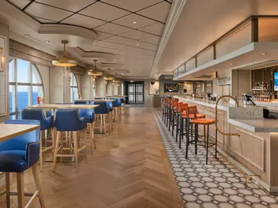 Innenansicht der Bar "The Local" an Bord der Norwegian Spirit. Im Vordergrund Tische mit blauen Hockern und dahinter eine lange Bar mit orangen Hockern. Große Fenster bieten Meerblick.