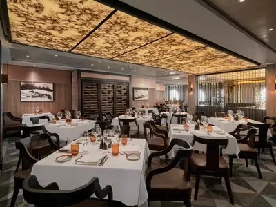Ein elegantes Spezialitätenrestaurant an Bord der Norwegian Spirit mit vielen leeren Tischen, weißer Tischwäsche und dunklen Holzstühlen. Im Hintergrund sind ein großes Weinregal und moderne Wandkunst zu sehen. Die Decke ist mit einer beleuchteten, marmorierten Textur versehen.