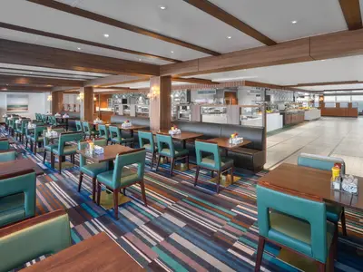 Geräumiges Garden Cafe Buffet-Restaurant an Bord der Norwegian Spirit. Viele Tische, türkisfarbene Stühle, dunkelbraune Bänke auf buntem Teppich. Essensstationen und Fenster mit Meerblick im Hintergrund.