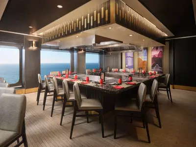 Elegantes Teppanyaki-Restaurant der Norwegian Spirit mit Meerblick. Lange Kochtheke, mit roten Servietten und Geschirr gedeckt. Moderne Stühle säumen die Tische.