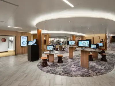Die moderne Foto-Galerie an Bord der Norwegian Spirit, ausgestattet mit mehreren digitalen Touchscreens zur Bildauswahl und Ausstellungstischen. Elegante Holzlamellen und eine geschwungene Decke prägen den Raum. Ein Schild bewirbt 40% Rabatt auf Fotografien.