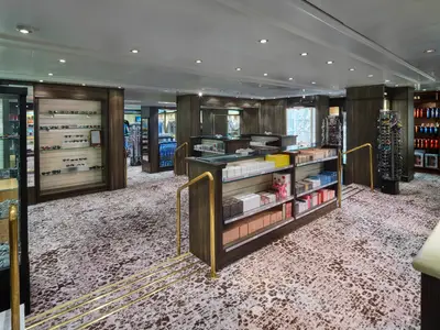 Heller Shop an Bord der Norwegian Spirit. Auslagen zeigen Sonnenbrillen, Parfüm und Produkte. Dunkle Holzregale, goldene Handläufe und gemusterter Teppich prägen das Interieur.