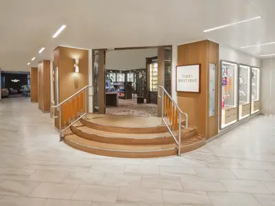 Ein heller Flur an Bord der Norwegian Spirit führt zu einer eleganten Treppe aus Holz, die den Eingang zur Tides Boutique markiert. Rechts sind beleuchtete Vitrinen mit Produkten zu sehen.