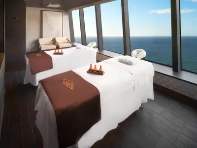 Ein luxuriöser Spa-Behandlungsraum an Bord der Norwegian Spirit mit zwei weiss bezogenen Massageliegen. Durch grosse Fenster ist der weite Ozean zu sehen. Auf den Liegen liegen braune Tücher und Massageöle.