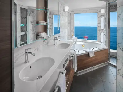 Ein geräumiges, modernes Badezimmer an Bord der Norwegian Spirit mit Doppelwaschbecken, großer Badewanne und Meerblick. Champagnerglas und Blumen sind zu sehen.