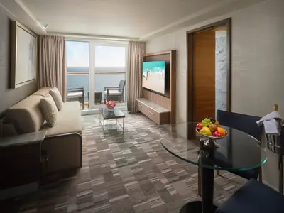 Blick in den eleganten Wohnbereich einer Penthouse Suite an Bord der Norwegian Spirit mit Sofa, Couchtisch und Zugang zum Balkon mit Meerblick. Ein Serviertisch mit frischem Obst und Champagnerflasche steht bereit.