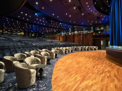 Innenansicht des Stardust Theaters an Bord der Norwegian Spirit. Gestufte Reihen mit blauen Sitzgelegenheiten, dahinter beige Sessel mit Tischen, und eine große Holzbühne.