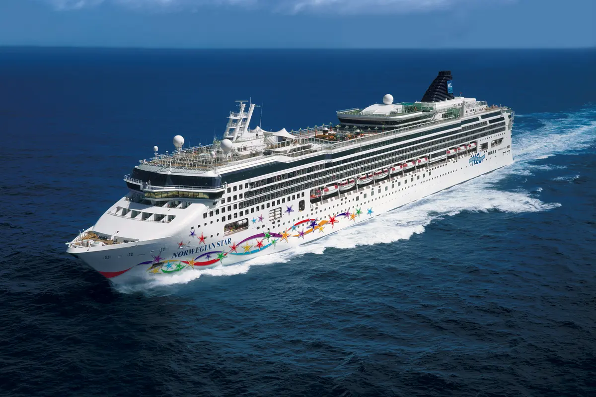 Luftaufnahme der Norwegian Star, einem weißen Kreuzfahrtschiff mit bunter Sternenbemalung am Bug, das auf dem blauen Meer fährt und eine weiße Heckwelle hinterlässt. Klarer blauer Himmel.