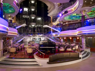 Das mehrgeschossige Atrium der Norwegian Star zeigt eine elegant beleuchtete Lounge mit Klavier, Bar, Tischen, Sofas, einer breiten Treppe und gläsernen Aufzügen unter einem Buntglasdach.
