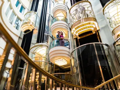 Ein Paar lächelt und blickt nach vorne, während es auf einem gläsernen Balkon im lichtdurchfluteten Atrium der Norwegian Star steht. Rundherum sind weitere Balkone und schimmernde Glasaufzüge sichtbar, die eine moderne und elegante Atmosphäre schaffen.