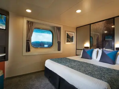 Geräumige Aussenkabine an Bord der Norwegian Star mit komfortablem Doppelbett, blauen Kissen und einem Bullaugenfenster mit Blick auf das blaue Meer. Ein Fernseher ist links zu sehen.