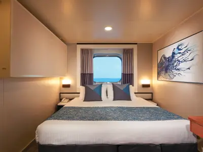Helle, moderne Außenkabine an Bord der Norwegian Star mit Doppelbett, Nachttischen und Bullauge mit Meerblick. Ein blaues Kunstwerk schmückt die Wand.