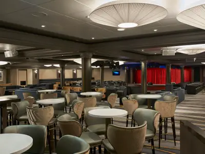 Der elegante Bar-Lounge-Bereich an Bord der Norwegian Star ist mit vielen Tischen, modernen Stühlen und einer beleuchteten Bühne mit roten Vorhängen ausgestattet. Gedämpftes Licht schafft eine intime Atmosphäre.