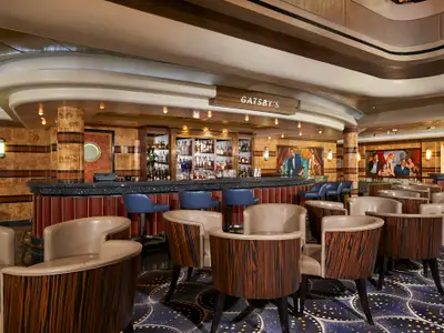 Ein Blick in die Gatsby's Bar an Bord der Norwegian Star. Man sieht die dunkle Bartheke mit Barhockern und beige-braune Sessel in einem eleganten Interieur im Art-Déco-Stil.