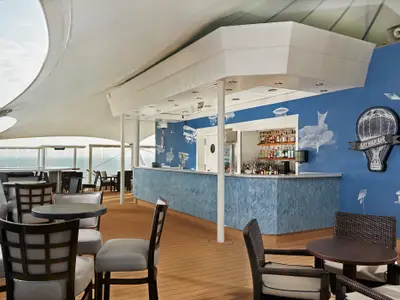 Die Sky High Bar an Bord der Norwegian Star, eine offene Bar auf dem Oberdeck. Blaue Wand mit Luftfahrzeug-Motiven und Heißluftballon-Logo. Gemütliche Sitzgruppen und Meerblick.