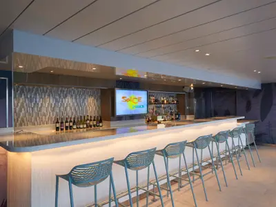 Blick auf die leere Spice H2O Bar an Bord der Norwegian Star. Ein langer, beleuchteter Bartresen mit türkisblauen Barhockern und Flaschenregalen ist zu sehen.