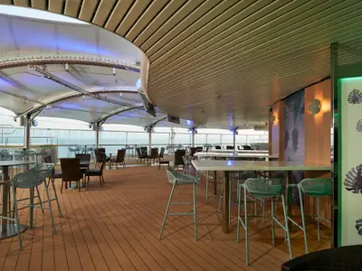 Ein heller, überdachter Bar-Lounge-Bereich an Deck der Norwegian Star. Reihen von Tischen und Stühlen stehen auf Holzplankenboden. Im Hintergrund ist das offene Meer zu sehen.