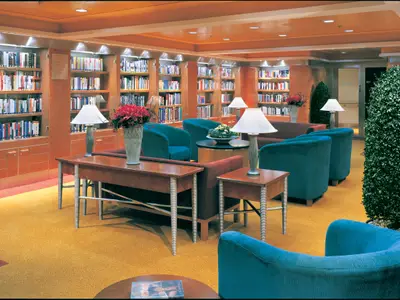Die Bibliothek der Norwegian Star. Gut beleuchtete Holzbücherregale mit Büchern, bequeme, tealfarbene Sessel und Beistelltische mit Lampen. Eine einladende Leseumgebung an Bord.