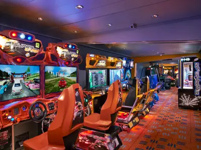 Die bunte Spielhalle der Norwegian Star zeigt eine Reihe von leuchtenden Rennsimulatoren und klassischen Arcade-Spielen auf einem gemusterten Teppichboden. Orangefarbene Sitze und Lenkräder laden zum Spielen ein.