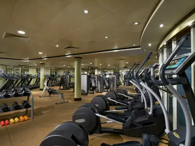 Modernes Fitnessstudio an Bord der Norwegian Star mit Reihen von Crosstrainern, Laufbaendern und Kraftgeraeten. Hantelregale und ein Rudergeraet im Vordergrund. Grosse Fensterfront mit Tageslicht. Ohne Personen.