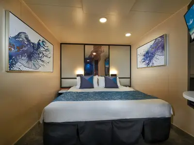 Helle Innenkabine der Norwegian Star. Großes Doppelbett mit blauer Tagesdecke und Kissen. Maritime Kunstwerke in Blautönen an den Wänden für eine moderne, einladende Atmosphäre.