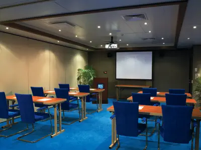Ein leerer, modern eingerichteter Konferenzraum an Bord der Norwegian Star mit blauen Stühlen, braunen Tischen und einer großen weißen Leinwand an der Stirnseite, bereit für Präsentationen.