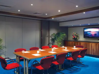 Ein heller Konferenzraum an Bord der Norwegian Star mit einem langen Holztisch, roten Stühlen und einem großen Flachbildfernseher an der Wand. Grüne Pflanzen dekorieren den Raum.