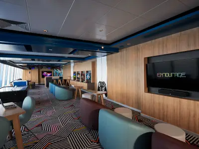 Blick in die moderne Entourage Lounge an Bord der Norwegian Star. Der Raum verfügt über mehrere Spielkonsolen, einen großen Fernseher mit Soundbar, Airhockey und bequeme Sitzgelegenheiten in verschiedenen Farben. Große Fenster bieten Meerblick.
