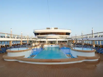 Pooldeck der Norwegian Star: Großer Pool, zwei Whirlpools und viele blaue Liegestühle. Die oberen, verglasten Decks des Schiffes sind im Hintergrund unter blauem Himmel zu sehen. Entspannte Atmosphäre.