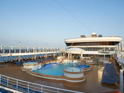 Das sonnige Pooldeck der Norwegian Star. Zwei große Pools und mehrere Whirlpools sind von einem Holzdeck und blauen Liegestühlen umgeben. Im Hintergrund das Meer, ein moderner Schiffsbau und klarer blauer Himmel. Die Szene ist menschenleer und ruhig.