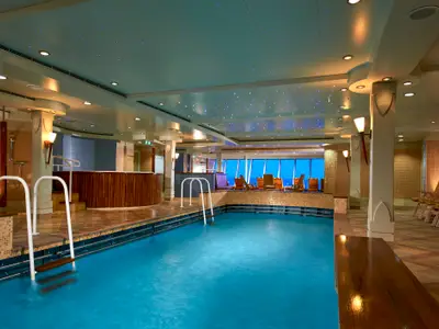 Innenpoolbereich an Bord der Norwegian Star mit blauem Wasser, einem runden Jacuzzi und Lounge-Stühlen vor Panoramafenstern zum Meer. Die Decke ist mit kleinen blauen Lichtern verziert.