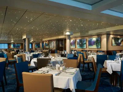 Ein elegantes Restaurant an Bord der Norwegian Star mit zahlreichen, weiß gedeckten Tischen. Die Bestuhlung ist abwechselnd in tiefem Blau und warmem Braun gehalten. Die Decke ist mit vielen kleinen Lichtern versehen, die einen Sternenhimmel simulieren. An den Wänden hängen moderne Kunstwerke.