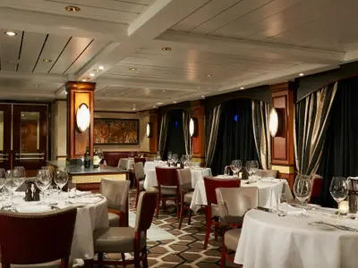 Elegantes Spezialitätenrestaurant an Bord der Norwegian Star. Festlich gedeckte Tische mit weißen Tischdecken, Gläsern und Besteck, rote und beige Lederstühle, dunkle Vorhänge.