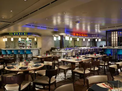 Innenansicht des modern gestalteten Spezialitätenrestaurants Ginza an Bord der Norwegian Star, mit einer beleuchteten Sushi-Bar, einer Sake-Bar mit blauen Hockern und zahlreichen Esstischen.