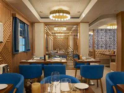 Innenansicht eines modernen Restaurants an Bord der Norwegian Star mit blauen Samtstühlen und dunklen Holztischen. Die Wände sind mit Holzpaneelen und teilweise mit blau-weißen Mustertapeten verziert. Eine goldene Ring-Deckenleuchte beleuchtet den Raum. Im Hintergrund ist ein abgetrennter Bereich mit weiteren Tischen und Sitzbänken zu sehen.