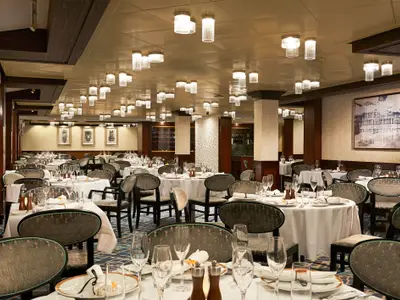 Elegantes Restaurant an Bord der Norwegian Star. Zahlreiche gedeckte Tische mit Gläsern, Besteck und stilvollen Stühlen. Moderne Deckenleuchten und Kunstwerke schaffen ein gediegenes Ambiente.