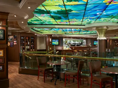 Innenansicht der O'Sheehan's Bar & Grill an Bord der Norwegian Star. Bunte Deckenverglasung, dunkle Holzeinrichtung, Tische und eine Bar schaffen eine gemütliche Pub-Atmosphäre.