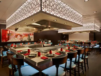 Das Teppanyaki Restaurant der Norwegian Star. Moderne Kochstationen mit blauen Stühlen und roten Gedecken. Geometrische Deckenleuchten und ein abstraktes Wandgemälde zieren den eleganten Raum.