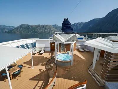 Sonniges Oberdeck der Norwegian Star mit rundem Whirlpool, Lounge-Sesseln und Essbereich. Im Hintergrund majestätische Berge und blaues Wasser.