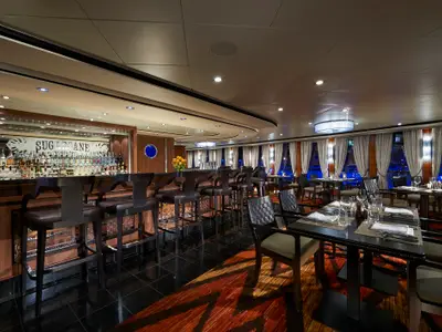 Blick in das elegant gestaltete Spezialitätenrestaurant Sugarcane an Bord der Norwegian Star. Eine Bar mit vielen Spirituosenflaschen ist im Vordergrund, dahinter mehrere gedeckte Tische.