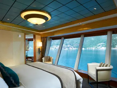 Luxuriöse Suite an Bord der Norwegian Star. Großes Bett, Sessel, Panoramafenster mit Blick auf bergige Küstenlandschaft und Stadt. Helle, elegante Einrichtung.