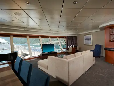 Innenansicht einer luxuriösen Suite an Bord der Norwegian Star. Ein großes Wohnzimmer mit Sofa, TV, Schreibtisch und einem Flügel bietet Panoramafenster mit Blick auf Meer und Küste.