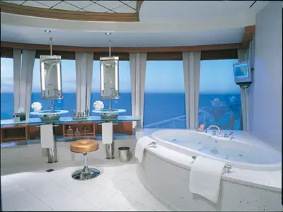 Ein elegantes Badezimmer an Bord der Norwegian Star mit Doppelwaschbecken, grosser Badewanne, weitem Meerblick und einem TV-Bildschirm an der Wand.