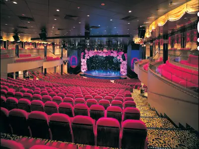 The Stardust Theater on the Norwegian Star, featuring multi-level seating with vibrant red velvet chairs. The stage is adorned with large pink flowers and sparkling lights. No people are visible.