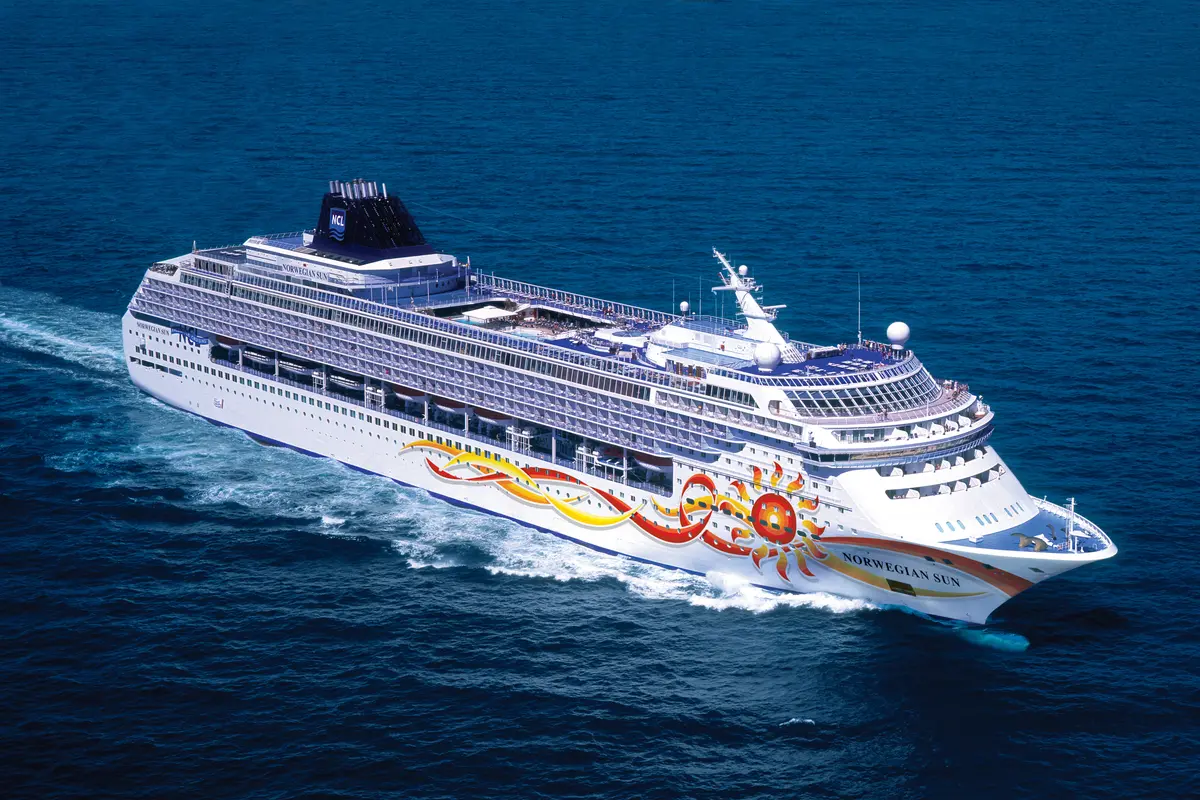 Luftaufnahme der Norwegian Sun auf offener See. Das weiße Kreuzfahrtschiff mit seiner markanten rot-gelben Sonnenbemalung am Bug hinterlässt eine Schaumspur im blauen Meer.
