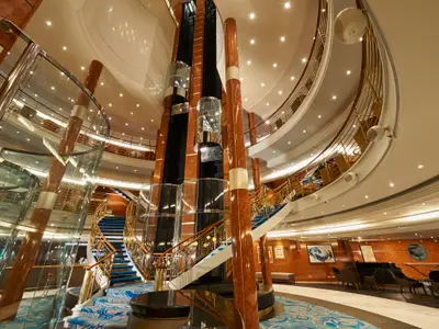 Weitwinkelansicht des mehrstöckigen Atriums an Bord der Norwegian Sun mit glänzenden Holzsäulen, Glasaufzügen, blauen Teppichtreppen und dekorativem Geländer.