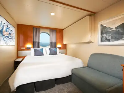 Eine gemütliche Aussenkabine der Norwegian Sun mit weißem Doppelbett, zwei Kunstwerken, einem blaugrauen Sofa und einem Bullauge mit Blick auf das Meer. Helle Holzvertäfelung und warme Beleuchtung tragen zur entspannten Atmosphäre bei.