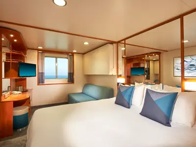 Geräumige Aussenkabine an Bord der Norwegian Sun mit grossem Doppelbett, einem Fenster mit Meerblick, einem Sofa und einem Schreibtisch mit TV. Modernes Design mit Holz- und Blautönen.