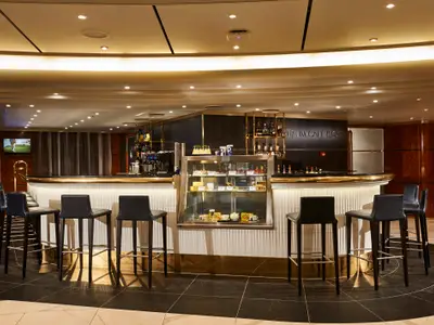 Innenansicht der Atrium Café & Bar an Bord der Norwegian Sun. Eine geschwungene Bar mit Barhockern und einer beleuchteten Vitrine ist sichtbar. Das Interieur ist elegant mit goldenen und dunklen Akzenten.